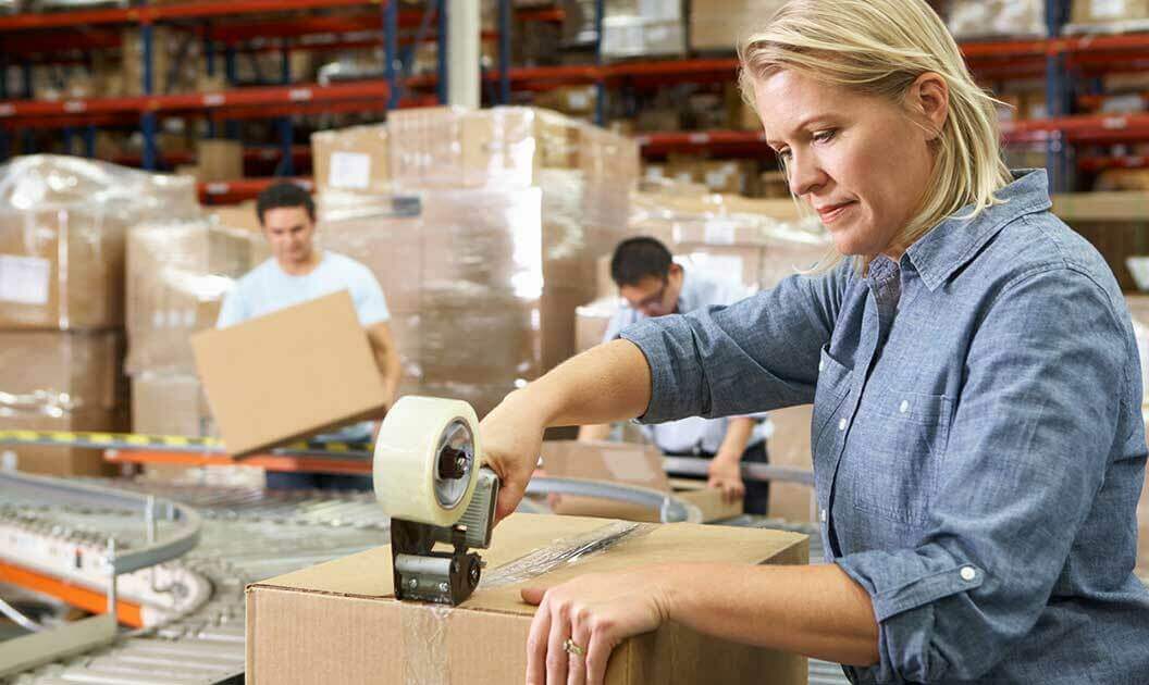 A warehouse worker taping up a box while her coworker lifts one off an assembly line behind her