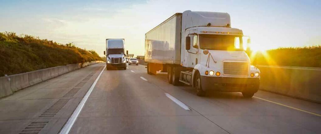 Two semis traveling on a highway near the end of the day